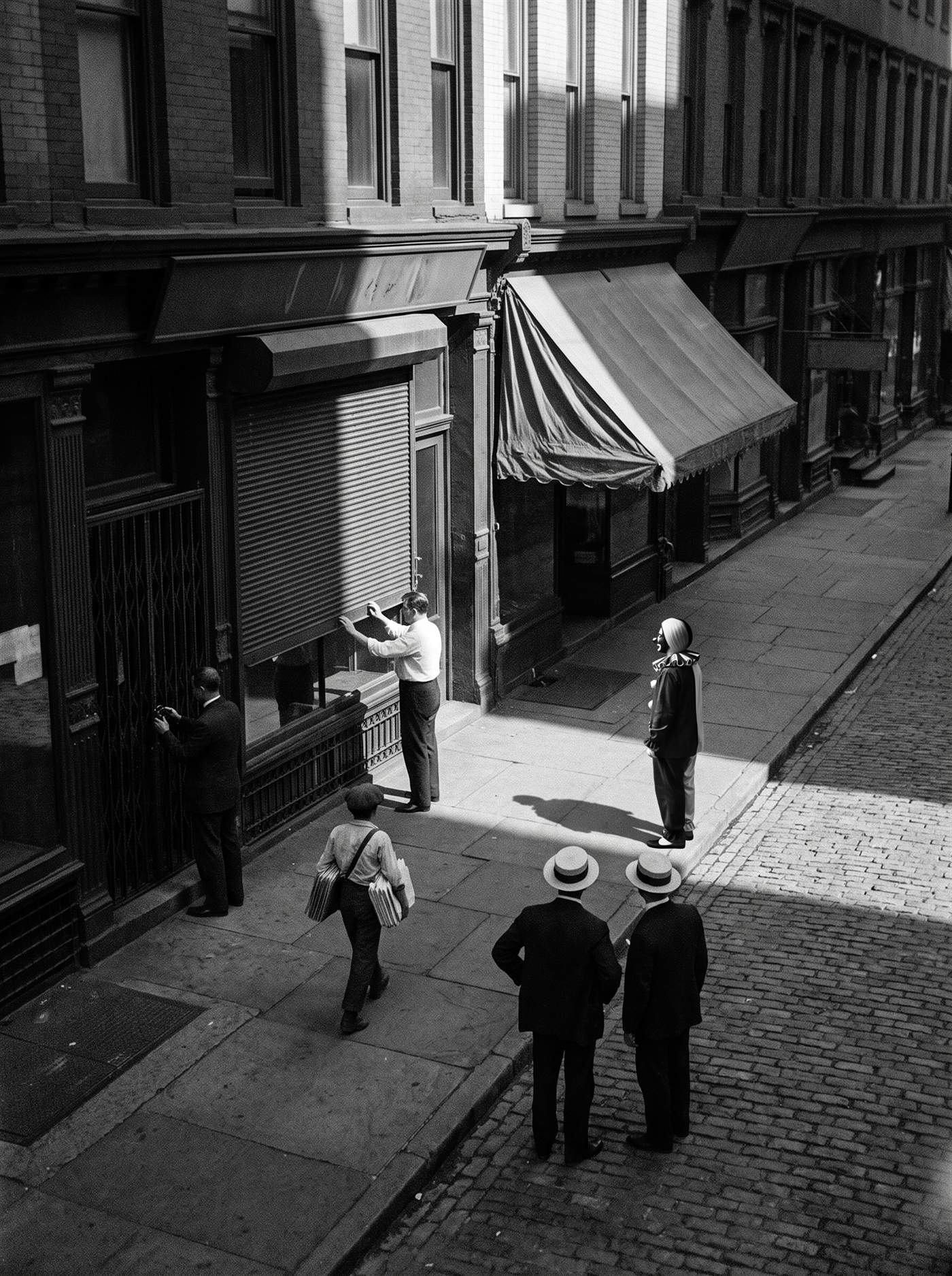 A monochrome press photograph looking down into a narrow lower Manhattan street from an upper window on a bright afternoon in July 1914. A clerk in shirtsleeves draws a folding shutter across a brokerage window, another man padlocks the grille of the doorway beside it, a newsboy walks away with a bundle under his arm, and two men in straw boaters stand at the kerb with their backs turned. The street is emptier than the hour warrants. Standing still in the one slab of sunlight on the pavement, small and casting a long shadow, is a clown whose face is split down the middle into one pale half and one dark half, his hair mirrored against it and a pale ruffled collar at his throat. Everyone else in the photograph is moving.