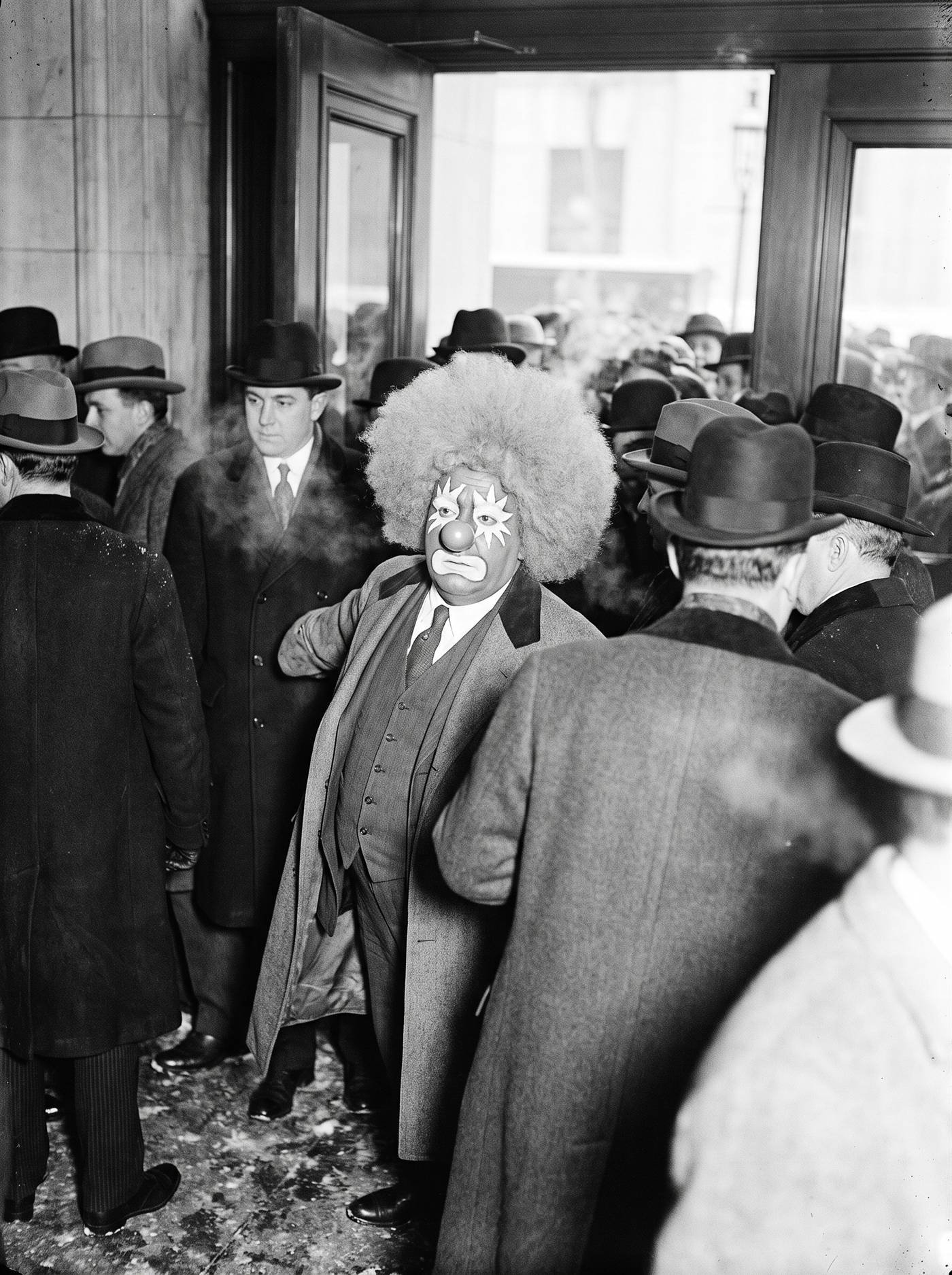 A monochrome press photograph taken with a hard flash inside the vestibule of the New York Stock Exchange in December 1914. A crowd presses in from the cold through the newly opened doors, overcoats buttoned to the throat, breath steaming, wet prints tracked across the stone floor. Among them, carried along by the crowd rather than leading it, is a heavy-set clown with a pale painted face, white starbursts radiating around both eyes, a bright ball nose and a huge round pale afro, his overcoat hanging open where everyone else's is fastened. His head is turned up and to the side toward the doors behind him.