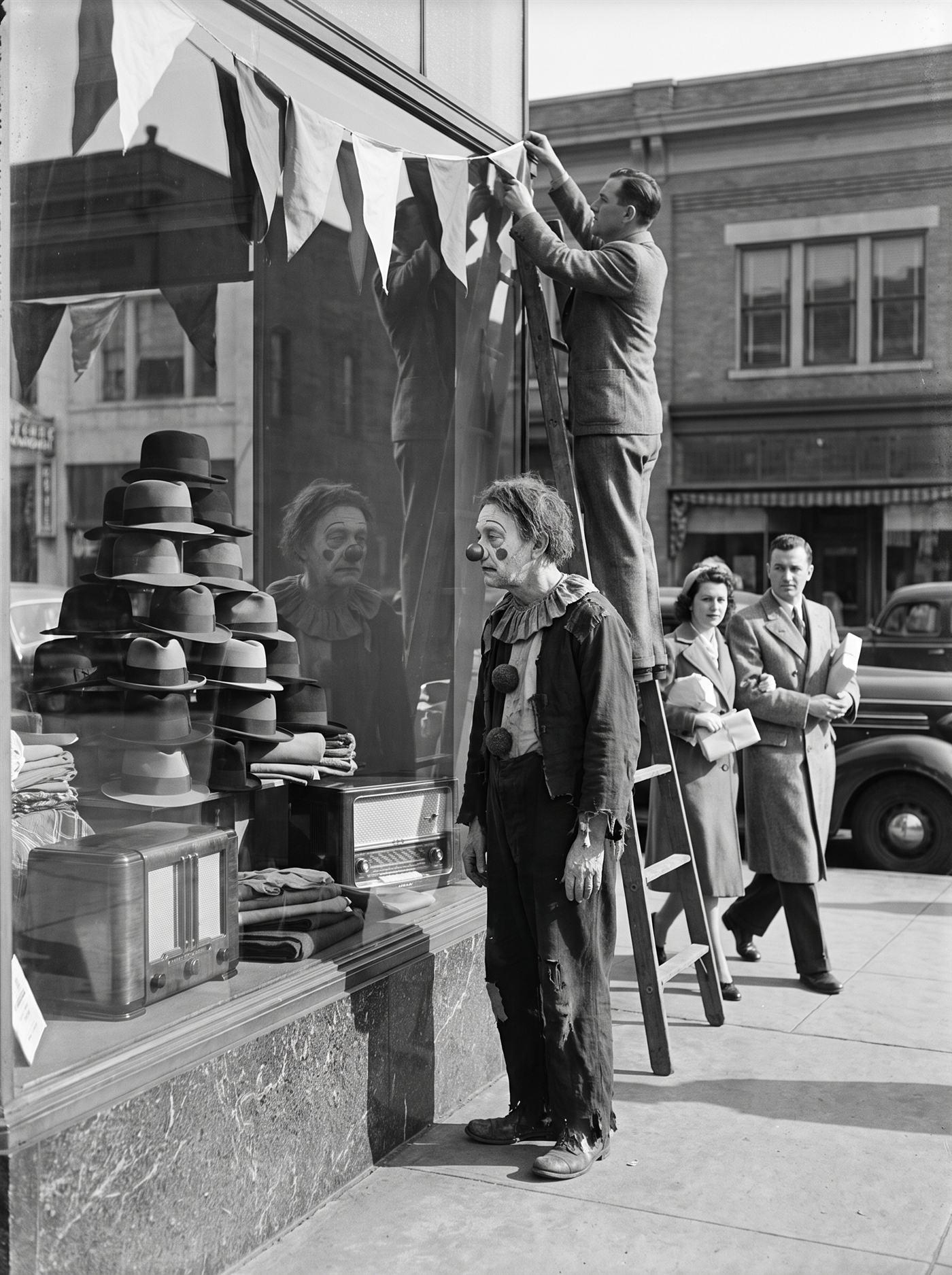 A black-and-white photograph of a freshly reopened main-street shopfront on a cold bright March morning in 1937. The plate-glass window is dressed with fine goods again — a pyramid of new hats, folded cloth, a gleaming radio cabinet — and a man on a ladder hangs fresh bunting above it while a well-dressed couple passes with parcels. Close to the glass, in profile, stands a gaunt old clown in a ruined ruff and patched coat, his cracked ash-white face and tarnished nose ghosted faintly in the window's reflection — his image hanging among the merchandise, between the hats and the radio, the one item the recovery did not restore. Nobody notices him.