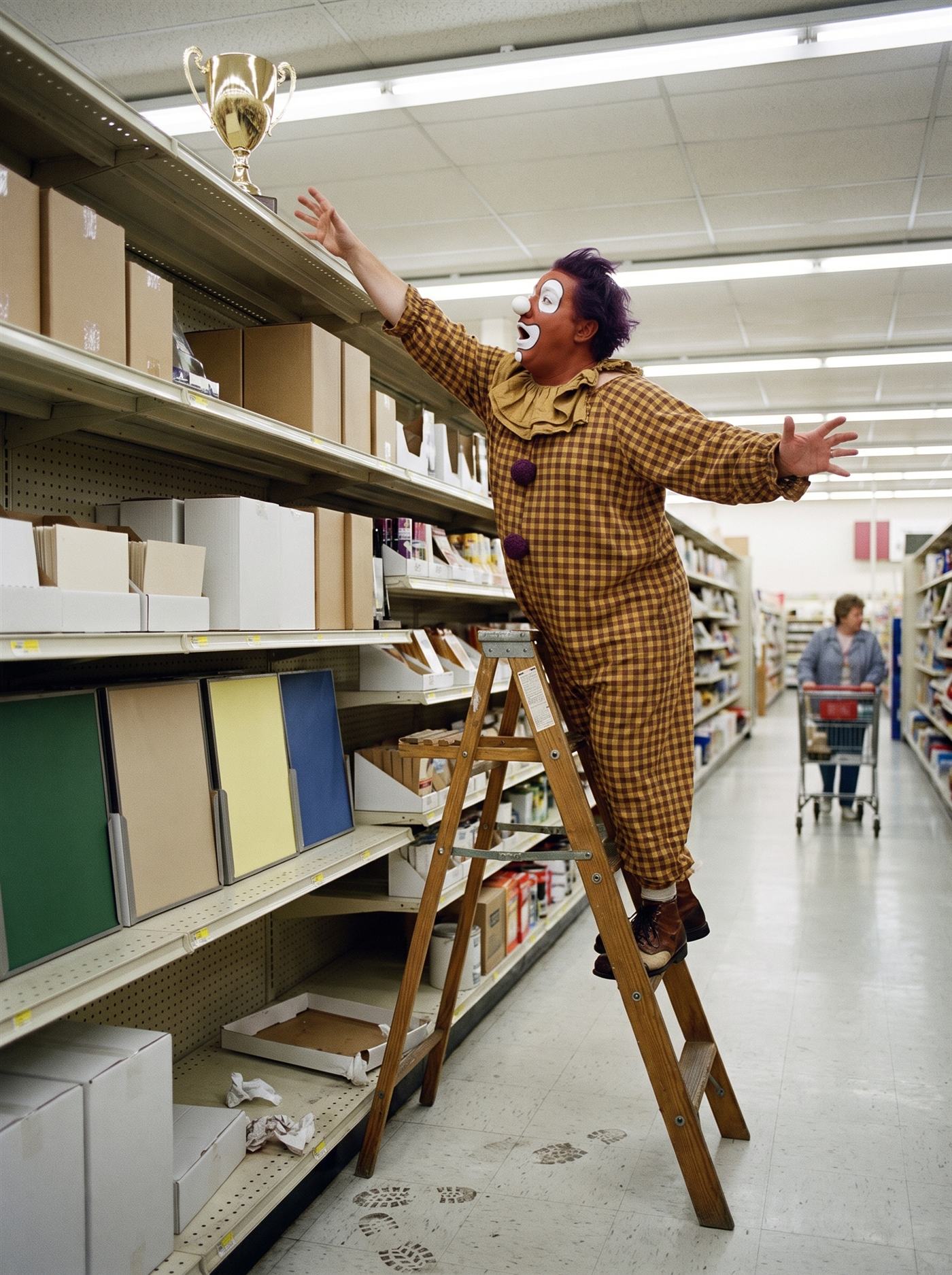 A color photograph inside a 1990 department store's tall-shelf aisle under flat fluorescent light. On the very top shelf, pushed to the back, sits a single gleaming unengraved golden trophy cup. A heavyset clown in a brown-and-mustard gingham costume with a torn mustard ruff, a terracotta-painted face with big mismatched white eye patches, an astonished open-O mouth and a wild purple mop of hair stands on the top step of a wooden stepladder at absolute full stretch — one arm extended to its limit, fingertips inches short of the trophy, his other arm windmilling, one foot lifting, the ladder beginning the faintest tilt. The exact instant between reaching and falling, held. Down the aisle a shopper pushes her cart past without a glance.
