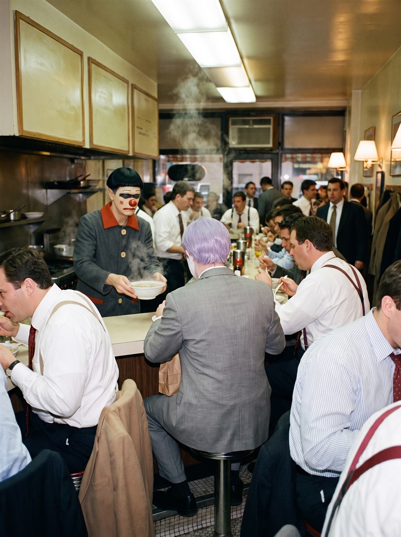 A color photograph of a small noodle counter in downtown New York at the peak of the busiest lunch rush it has ever had, autumn 1997. Every stool is taken by office workers in shirtsleeves, loosened ties and suspenders, trench coats over stool backs, more customers standing to eat, steam rising off the bowls. Behind the counter, calm in the middle of the crush, a lean clown in an ivory-painted face with a single dark band brushed across both eyes, a glossy red nose, a flat level red mouth and a jet-black bowl-cut wig sets a steaming bowl down with one hand — the one unhurried figure in the room. At mid-counter, seen entirely from behind, a man in a too-big grey suit and tidy lavender wig eats like everyone else, a small paper bag resting against his hip.