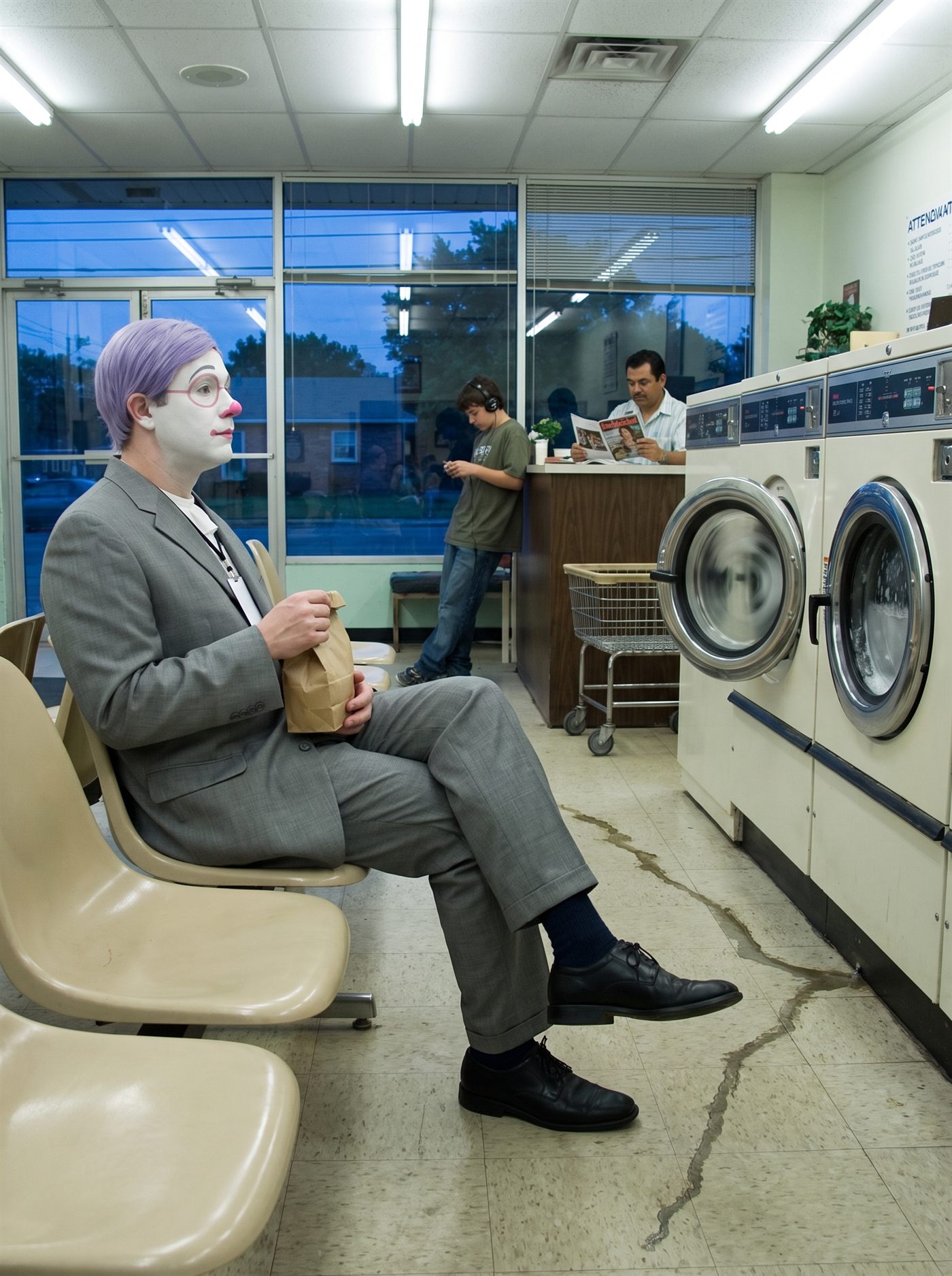 A slightly clinical early-DSLR photograph of a neighborhood laundromat on a summer evening in 2007, fluorescent panels against the last blue of dusk in the storefront glass. A man in full clown makeup — clean white face, painted rose spectacle rings, a lavender side-parted wig, a too-big grey suit with a blank badge on a lanyard — sits in profile on a molded chair, legs crossed, a small brown paper bag held closed in both hands on his lap, patient and content. In front of him one washer spins in a soft blur — and its neighbor stands stopped mid-cycle, the porthole a still circle of grey suds. A thin dark line runs along the linoleum from the back of the room toward the machines, catching the light. Nobody has seen it: not the clown, not the attendant reading at the counter, not the teenager slouched at the counter in headphones. The room goes on waiting for its laundry.