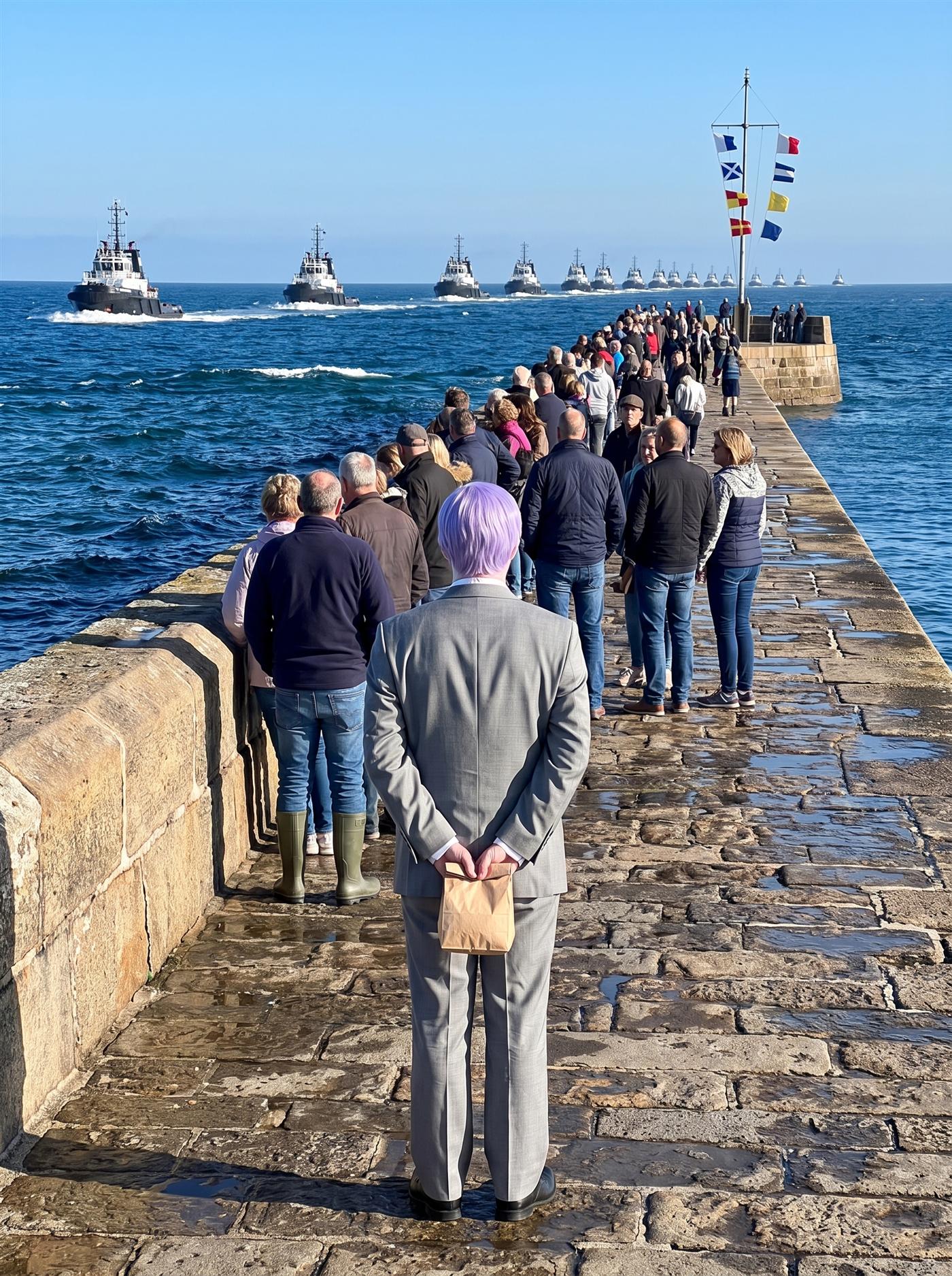 A photograph of a long stone breakwater at a harbor mouth on a bright hard morning after a week of storms, the sea a deep saturated ultramarine with white crests still running. On the horizon a fleet is arriving in force — a long line of large dark ships coming in abreast, bow waves white, more cresting behind them. A crowd has come out along the breakwater to watch: fishermen, shopkeepers, families, someone with binoculars, all facing out to sea, some pointing, more stunned than joyful. Among them at the center of the near ground, seen from behind, stands a man in full clown makeup and a grey suit a half size too big — tidy lavender side-parted hair — perfectly still amid the pointing crowd, his hands clasped behind his back holding a small brown paper bag.