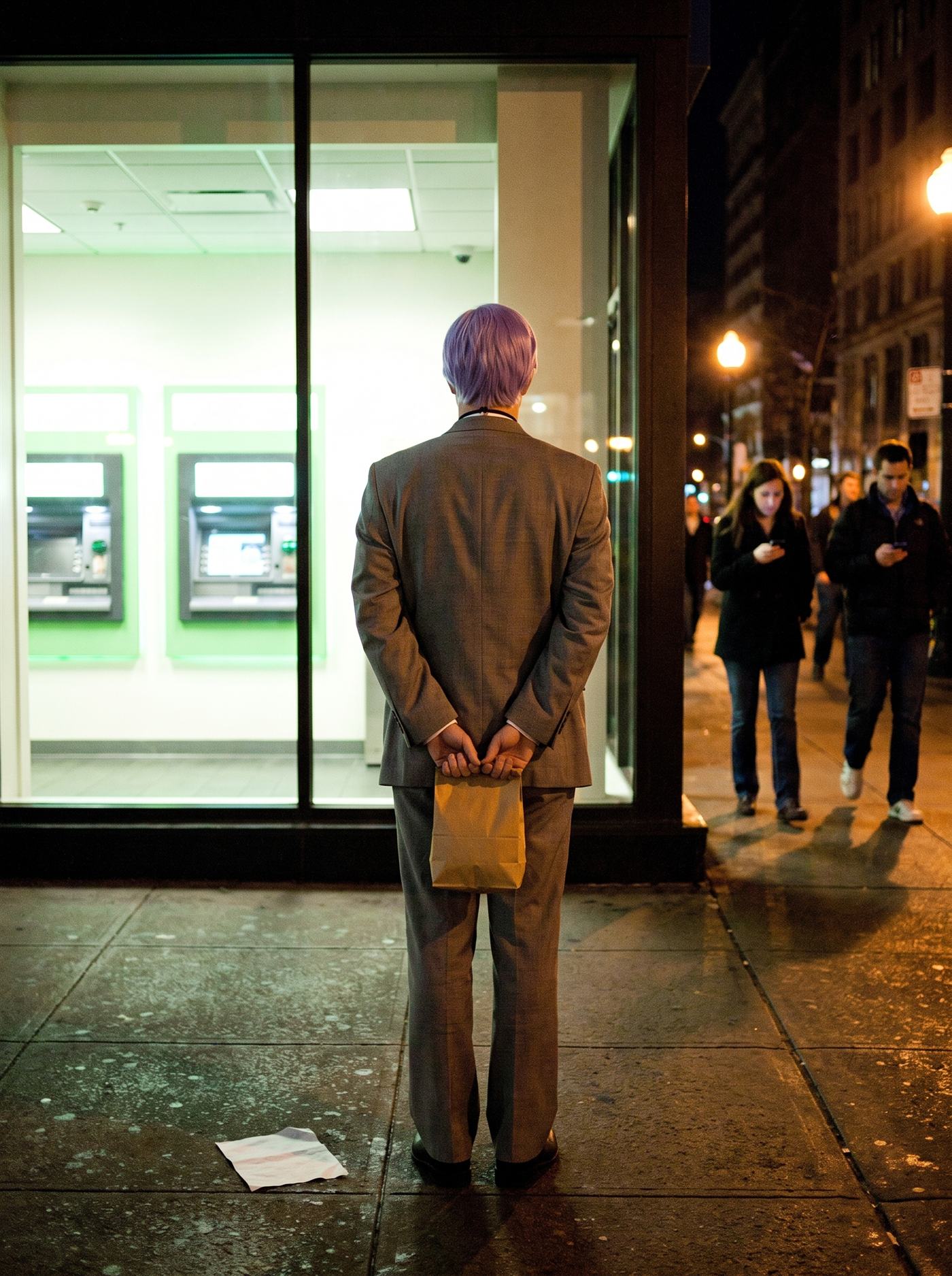 A color photograph of a bank branch's glass vestibule on a city street at night in August 2011 — the small ATM lobby glowing white-green through floor-to-ceiling glass, empty except for its machines. On the dark wet sidewalk just outside the glass stands a man in a too-big grey suit and a tidy lavender side-parted wig, seen entirely from behind, perfectly still, his hands clasped behind his back holding a small brown paper bag closed — looking into the lit empty lobby the way a man looks at something he has decided to trust one more time. A blank slip of paper lies at his feet. Behind him, passersby drift through the lamplight lit by their phones, paying no attention.