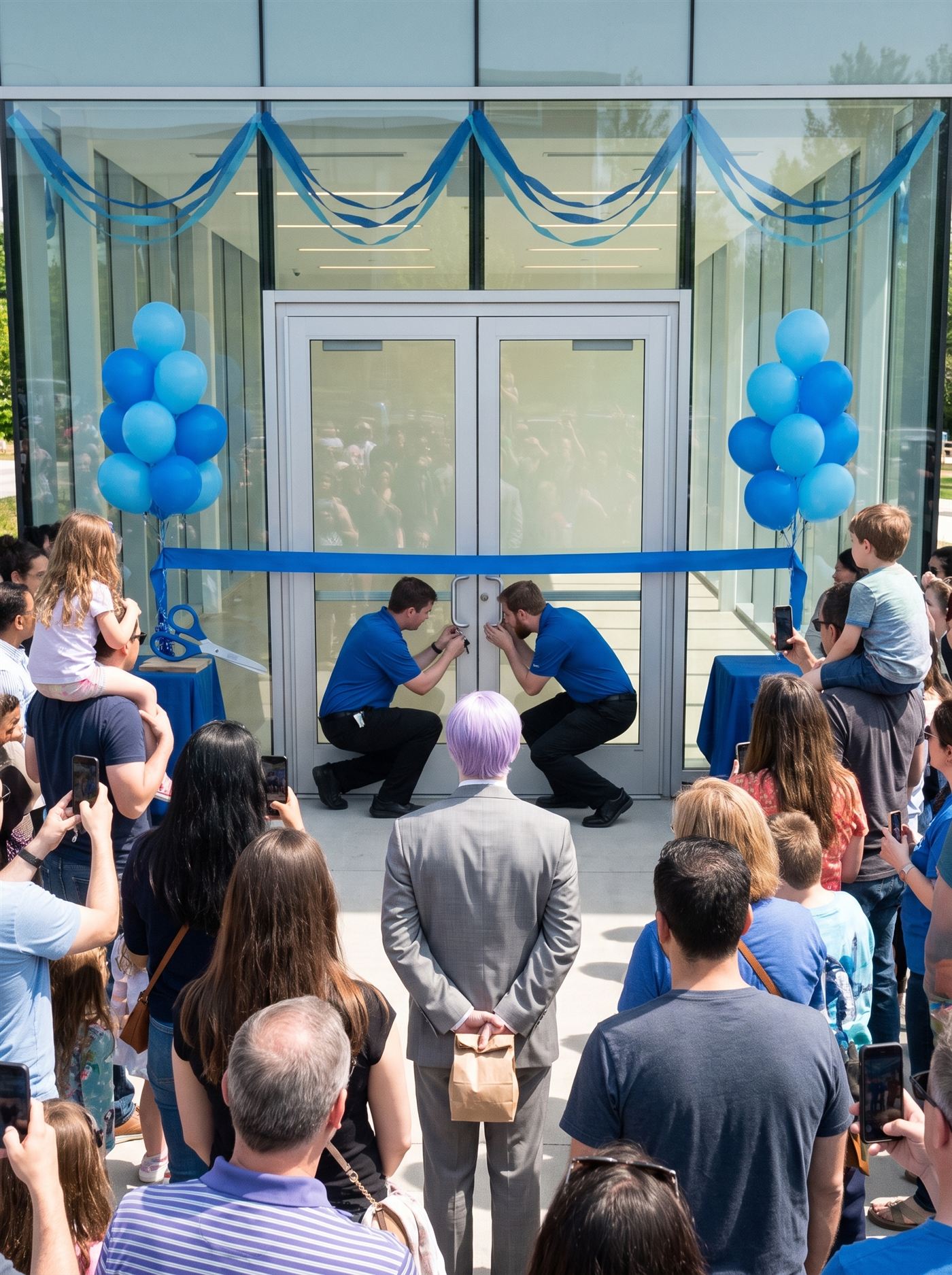 A photograph of the grand opening of a new glass-fronted pavilion on a bright late-morning in May, decked entirely in one shade of medium blue: clusters of blue balloons at the doorposts, blue bunting swagged across the facade, a wide blue ribbon stretched uncut across the double glass doors, oversized ceremonial scissors waiting on a small table. The crowd is far larger than anyone planned — pressed close, phones raised, kids on shoulders. At the doors, two staff in matching blue polos crouch at the lock, one working a ring of keys, the other peering in; the doors are not opening. The glass gives back the waiting crowd's own reflection over an interior that is lit, ready, and completely empty. In the middle of the waiting crowd, seen from behind, stands a man in full clown makeup and a grey suit a half size too big — tidy lavender side-parted hair — perfectly still, facing the ribboned doors, his hands clasped behind his back holding a small brown paper bag. Not one person looks at him.