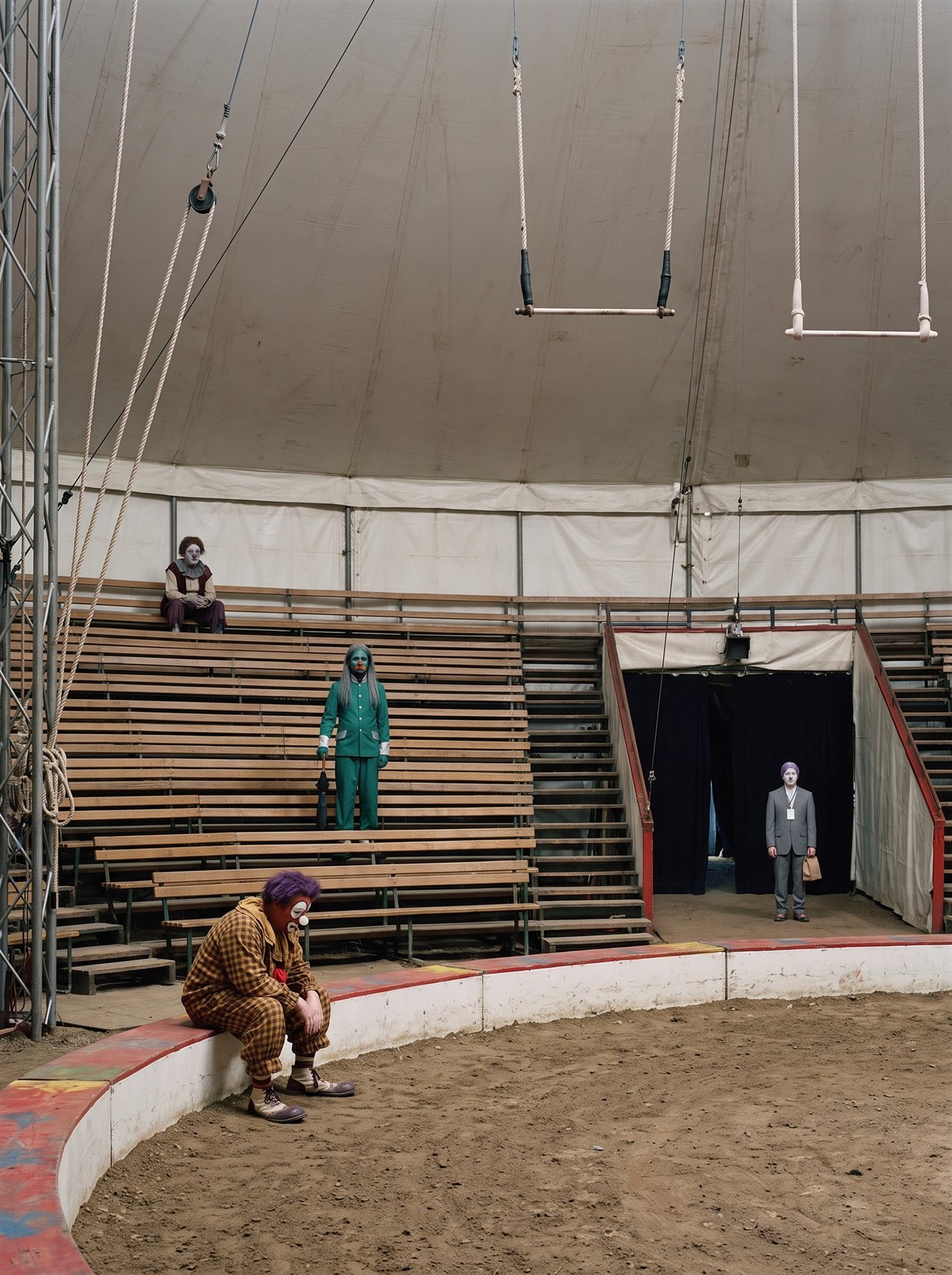 A photograph of the inside of a one-ring circus big top with the house lights on and every seat empty, March 2020. Trapezes hang still over the raked ring. On the ring curb sits a heavyset clown in terracotta face paint and brown-and-mustard gingham, elbows on knees, head bowed. Halfway up the empty grandstand a lean clown in jade-green greasepaint and a dark buttoned suit stands between the rows with a closed umbrella planted like a cane, looking out across the seats as if counting them. High at the top of the stands a gaunt clown with a cracked ash-white face sits with his hands folded. Far across the ring, at the mouth of the performers' tunnel, small in the shadow, stands a man in a grey suit with a white-painted face, a lanyard, and a small brown paper bag. Nobody performs. There is nobody to perform for.