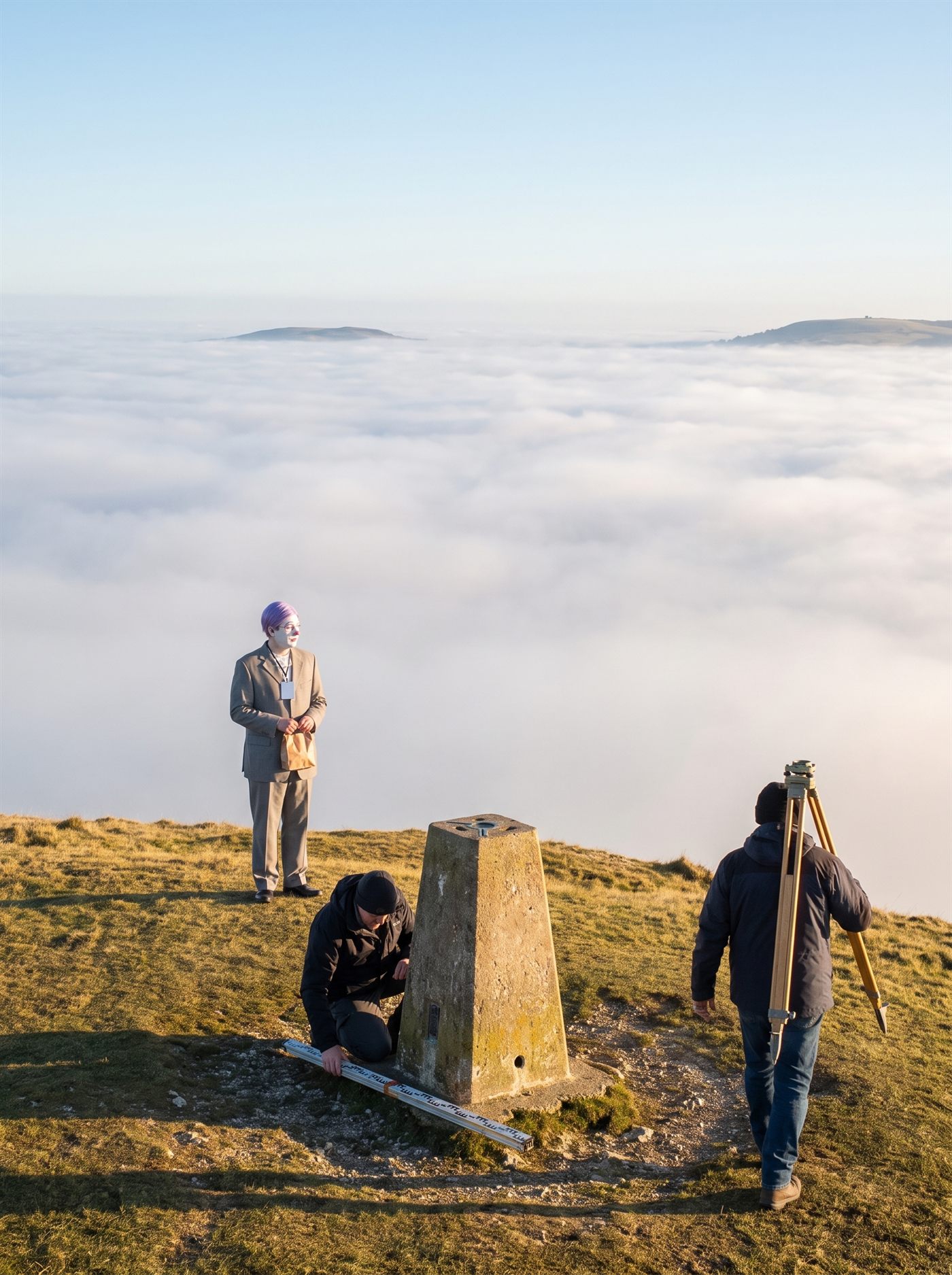 A photograph of the summit of a bare grass hill on a clear, cold morning in late January 2026. A squat weathered concrete triangulation pillar stands near the centre of the frame with a metal fitting set in its crown, and a surveyor in a dark jacket and cap kneels at its base beside a folded levelling staff laid on the turf. To the right a second surveyor walks away downhill with a yellow wooden tripod over his shoulder, seen from behind. Beyond and far below, a flat white sea of fog fills the entire valley and has swallowed everything in it; only two distant hill shoulders break the surface, and the pale sky above is empty. Low sun rakes across the summit grass and turns it gold, while everything below the fog line is gone. A man in a grey suit a half size too big stands alone on the left of the summit, small in the frame, seen mostly from behind and turned slightly toward the camera: white greasepaint over his face, rose-pink rings painted round his eyes, a glossy pink nose, a tidy lavender wig, a blank white badge on a lanyard. Both hands hold a single small brown paper bag at the front of his waist. He is looking out over the fog. Neither surveyor is looking at him.