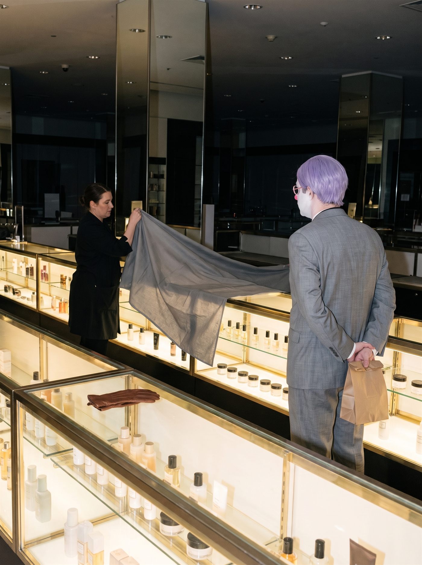 A photograph of a department store beauty hall at closing time, seen down the length of the counters. Long glass display cases lit warmly from within run away into the frame, their tops crowded with rows of small bottles and pots; tall mirrored columns above them double the emptiness of the hall, and the far end is already dark. In the left middle distance a saleswoman in a black smock lifts a large grey dust sheet and draws it out across one of the lit counters. A single brown leather glove has been left lying on the glass in the foreground. On the right, standing at the end of the counter run with his back to the camera, is a man in a grey suit a half size too big: a tidy lavender side-parted wig, white greasepaint at the cheek, a rose-pink painted eye ring just visible in profile. Both hands hold a single brown paper bag behind him at his lower back. He is watching the sheet come across. She has not looked up.