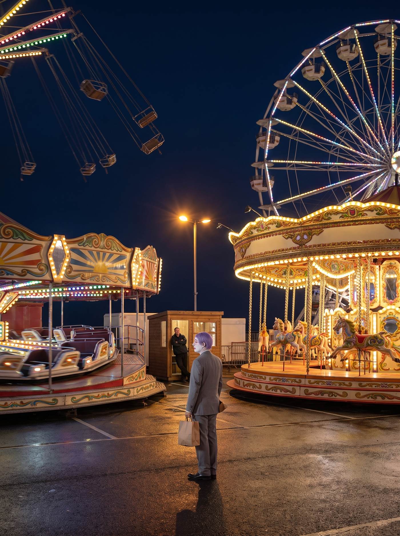 A photograph of a seaside funfair at night on wet black asphalt. Four rides are running and every one of them is empty: at the upper left a set of swing chairs flies out at full extension with nothing in the seats, motion-blurred against a deep blue sky; below them a waltzer-style ride spins, its cars empty and smeared with movement; on the right a large illuminated wheel turns with every gondola empty; and in the right foreground a carousel revolves with all its horses riderless, lit gold from the inside. Every ride carries thousands of bulbs and its fascia is painted in abstract scrollwork and sunbursts with no lettering anywhere. There is no crowd at all; the wide foreground is bare wet tarmac reflecting the lights. A single operator in a dark jacket leans in the doorway of a small wooden booth with his arms folded, looking at nothing. Alone in the middle of the empty ground, a man in a grey suit a half size too big stands quite still while everything around him moves, seen from behind and turned to look off to one side: a tidy lavender wig, white greasepaint, a rose-pink painted eye ring in profile, a single brown paper bag hanging from one hand at his side. The operator has not looked at him.
