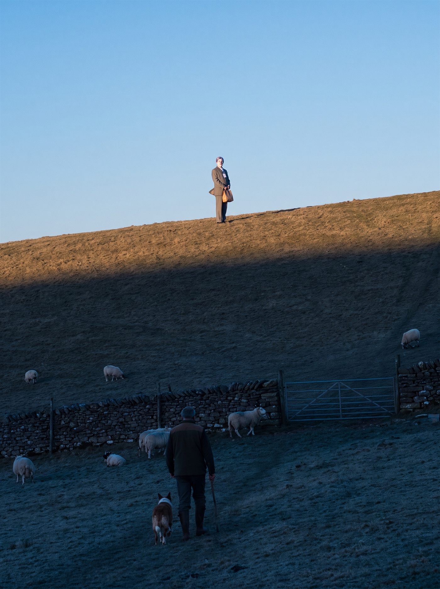 A photograph of a bare grass hillside at the very last of the light on a March evening. A hard, clean shadow line runs diagonally across the whole slope: above it a narrow strip of hilltop is still lit warm gold by the low sun, and below it the entire field has gone cold blue — a dry stone wall, a metal field gate and half a dozen grazing sheep all already in shadow. The sky above is pale and empty. A farmer in a dark coat walks downhill into the shadow with a stick in his hand and a collie at his heel, seen from behind. Alone on the skyline, very small in the frame and standing in the last lit strip, is a man in an off-the-rack grey suit with a tidy lavender wig and a white-painted face, a blank badge on a lanyard, holding a single small brown paper bag at the front of his waist. The shadow line is a few feet below his shoes and moving up the slope toward him. Nobody is looking at him.