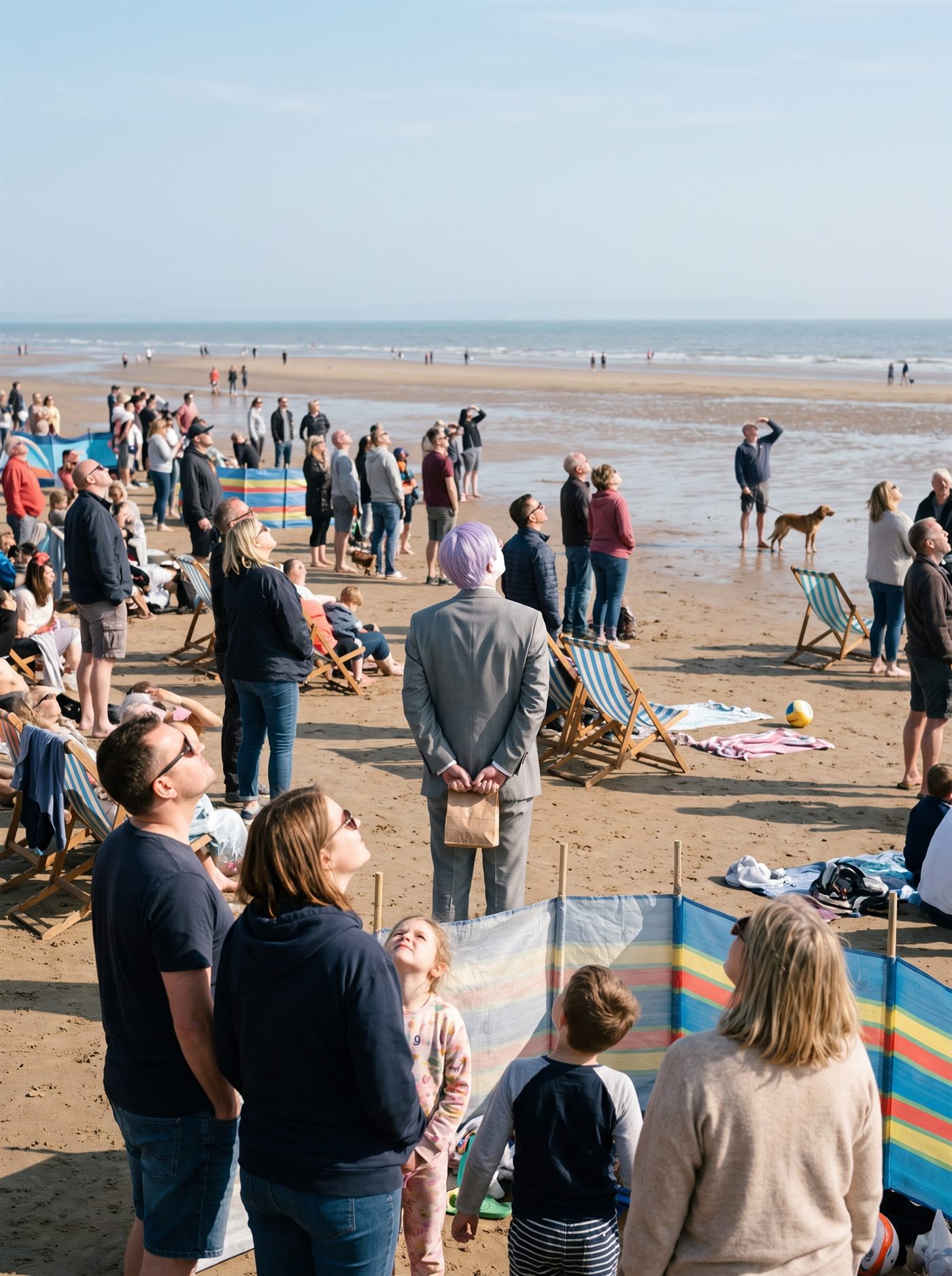 A photograph of a wide sandy beach on a bright cool afternoon during a flypast. Several hundred people are strung out across the sand in loose groups — families in deckchairs, a man with a dog at the water's edge, women in sun hats shielding their eyes — and every single one of them has stopped and tilted their head back to look up and to the right at the same point in the sky. The thing they are looking at is above the top edge of the frame and is never shown. Striped windbreaks and abandoned deckchairs sit at odd angles, a beach ball lies unattended on the wet sand, and the sea runs flat and pale along the far horizon. Standing among them in the middle distance, small in the frame and partly blocked by a striped windbreak and a family in the foreground, is a man in an off-the-rack mid-grey suit with tidy lavender side-parted hair, seen from directly behind, his head tipped back at the same angle as everyone else's, holding a small folded brown paper bag in both hands behind his back. Nobody is looking at him.