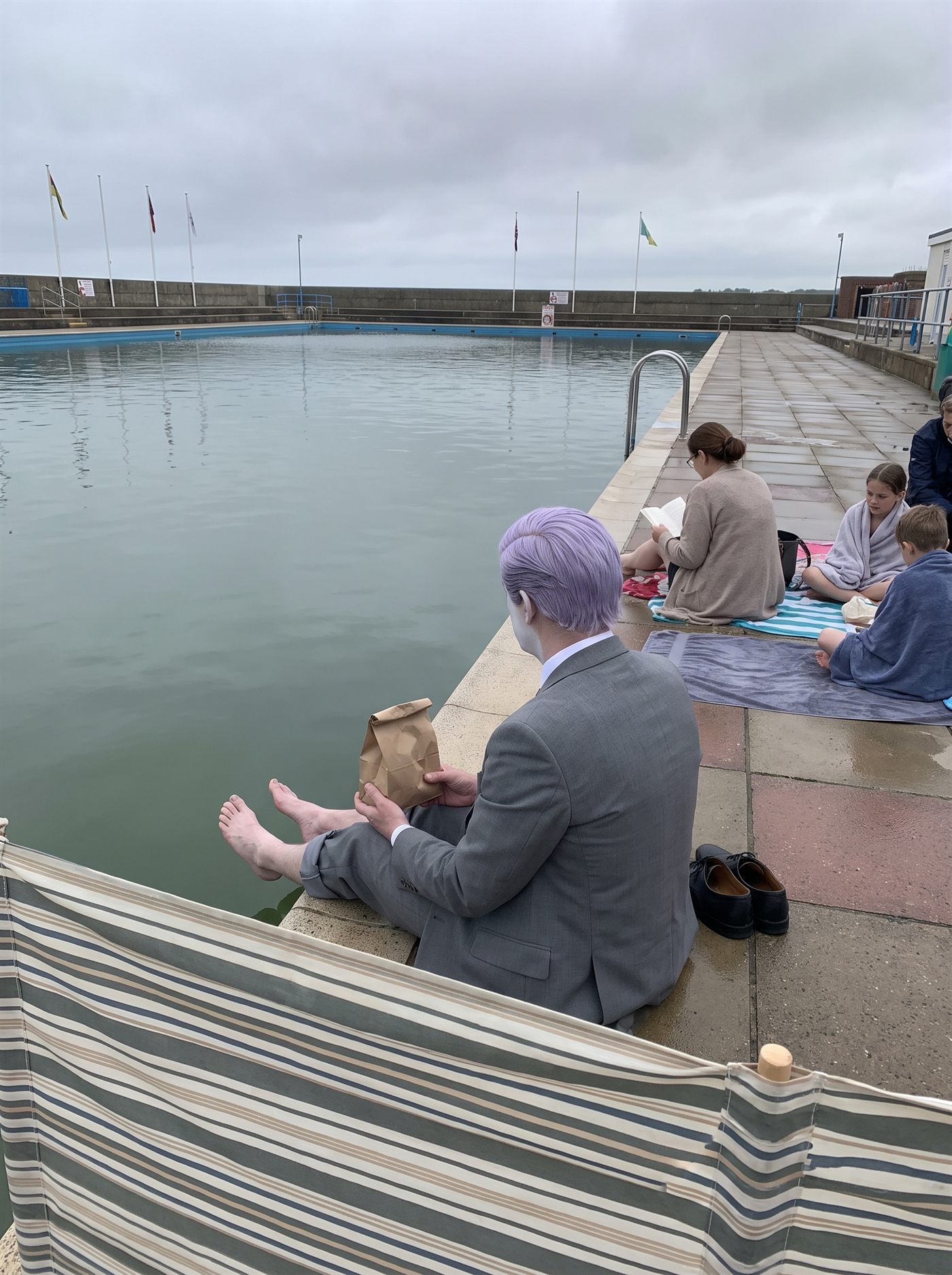 A photograph of a municipal open-air swimming pool under a completely flat, fully overcast sky. The pool fills most of the frame, its water a still, hard, pale grey-green with no one in it and no ripple on it. Along the far side, people sit out on the concrete in coats and cardigans pulled on over swimming things, one woman reading, two children wrapped in towels doing nothing; a row of flags on poles at the far end hangs straight down. In the near foreground a striped canvas windbreak has been set up against no wind at all. Sitting on the tiled edge of the pool, seen from directly behind, is a man in an off-the-rack mid-grey suit with tidy lavender side-parted hair, his trouser legs rolled to the shin and his shoes placed neatly on the tiles beside him, holding a small brown paper bag in both hands. His bare feet hang over the water without touching it. There is not one shadow anywhere in the picture. Nobody is looking at him.