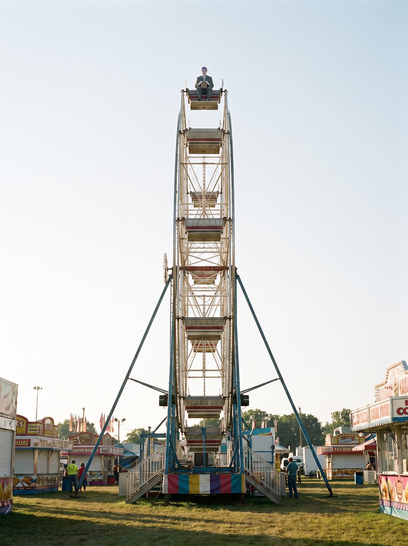 A photograph of a fairground on an early August morning, shot from the ground looking up: a tall Ferris wheel, stopped, against a pale thin morning-blue sky. The fair is barely awake — rides still, stalls shuttered, a worker far below on the empty midway. In the topmost gondola, small in the frame, sits a man in a grey suit with tidy lavender hair and a white-painted face — a tiny figure at the very apex, alone on the whole wheel, looking down over the safety bar at the ground far below, a small brown paper bag held in his lap. Every other gondola is empty. Nobody on the ground looks up.
