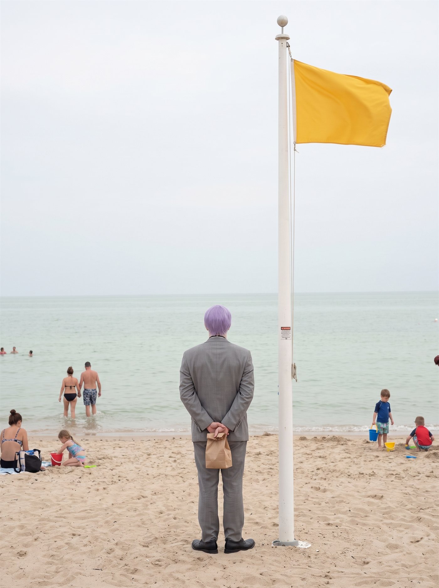 A photograph of a public beach on an overcast-bright August afternoon, the light flat and pearly. The sea is unusually calm — a long glassy swell in pale celadon green-grey, almost no surf — but at the lifeguard station's white flagpole a single solid yellow warning flag stands out stiff and straining in an offshore wind, the only thing in the frame that is not calm. Scattered beachgoers pay it no mind: a couple wading in, children digging with buckets, families on towels. Near the base of the flagpole, seen from behind, stands a man in full clown makeup and a grey suit a half size too big — tidy lavender side-parted hair — perfectly still, facing the flat water, his hands clasped behind his back holding a small brown paper bag closed behind him. He watches the calm water the way a man watches something he does not trust. Nobody is looking at him.