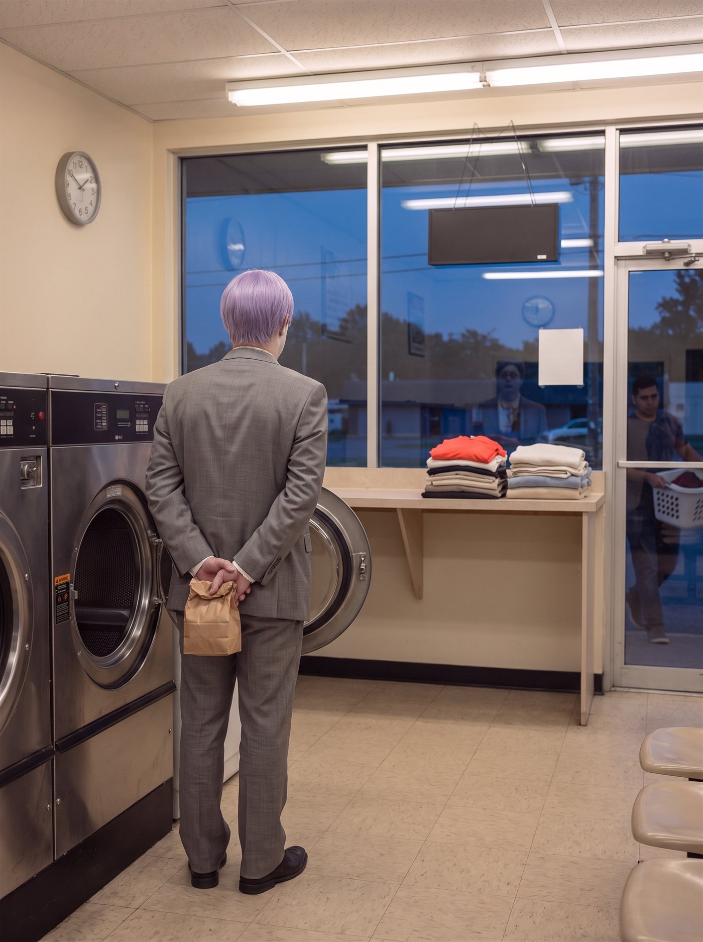 A photograph of the same small laundromat as the night before, one evening later: warm fluorescent light inside, deep blue dusk in the plate-glass windows, the plain round clock still on the bare wall. The row of steel front-loading machines stands quiet, and one of them has finished — its porthole door hangs wide open toward the room, the empty drum lit and still. A man in full clown makeup and a grey suit a half size too big stands in front of it, seen entirely from behind, feet together, head level, looking into the open drum. His hands are clasped behind his back, both holding a small brown paper bag closed. On the folding counter beside him the finished load is stacked in neat folded piles — white, beige, pale blue, grey — with one bright red-orange shirt folded on top, the only strong color in the room. At the far end, beyond the glass, a customer is leaving with a full laundry basket on their hip. Nobody is looking at him.