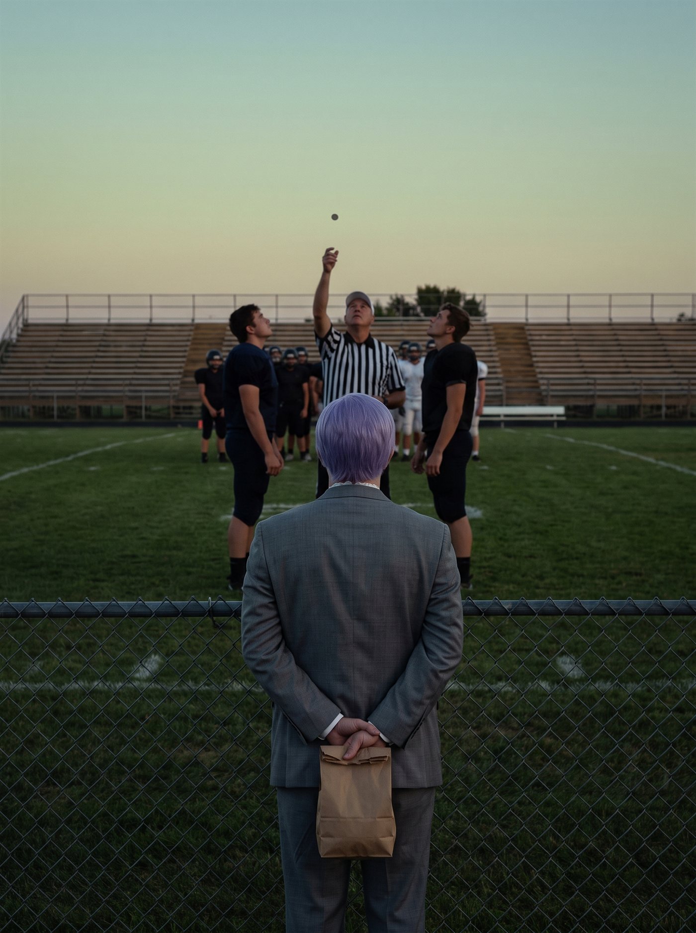 A photograph taken from behind a low chain-link fence at the sideline of a small-town football field at dusk. The sky is a pale, even green-gold going to peach at the horizon, and the field lamps on their poles are not lit, so the grass is a dim, flat green and nothing on it is lit harder than anything else. Beyond the fence, at midfield, a referee in a black-and-white striped shirt stands between two team captains in plain dark jerseys with his right arm raised, and a small coin hangs in the air above his hand; both captains have their heads tipped back watching it, and behind them the two teams stand in loose lines in front of a low set of empty metal bleachers. In the near foreground, on this side of the fence and a little left of center, a man in a grey suit a half size too big is seen squarely from behind, the back of his head to the camera, his head level and facing the field. His short lavender hair is combed flat above a white collar. His hands are clasped behind his back at the base of his spine, both fists closed around the rolled-over top of a plain paper bag with no handles, so that the bag hangs shut behind his legs. He is the only person on this side of the fence.