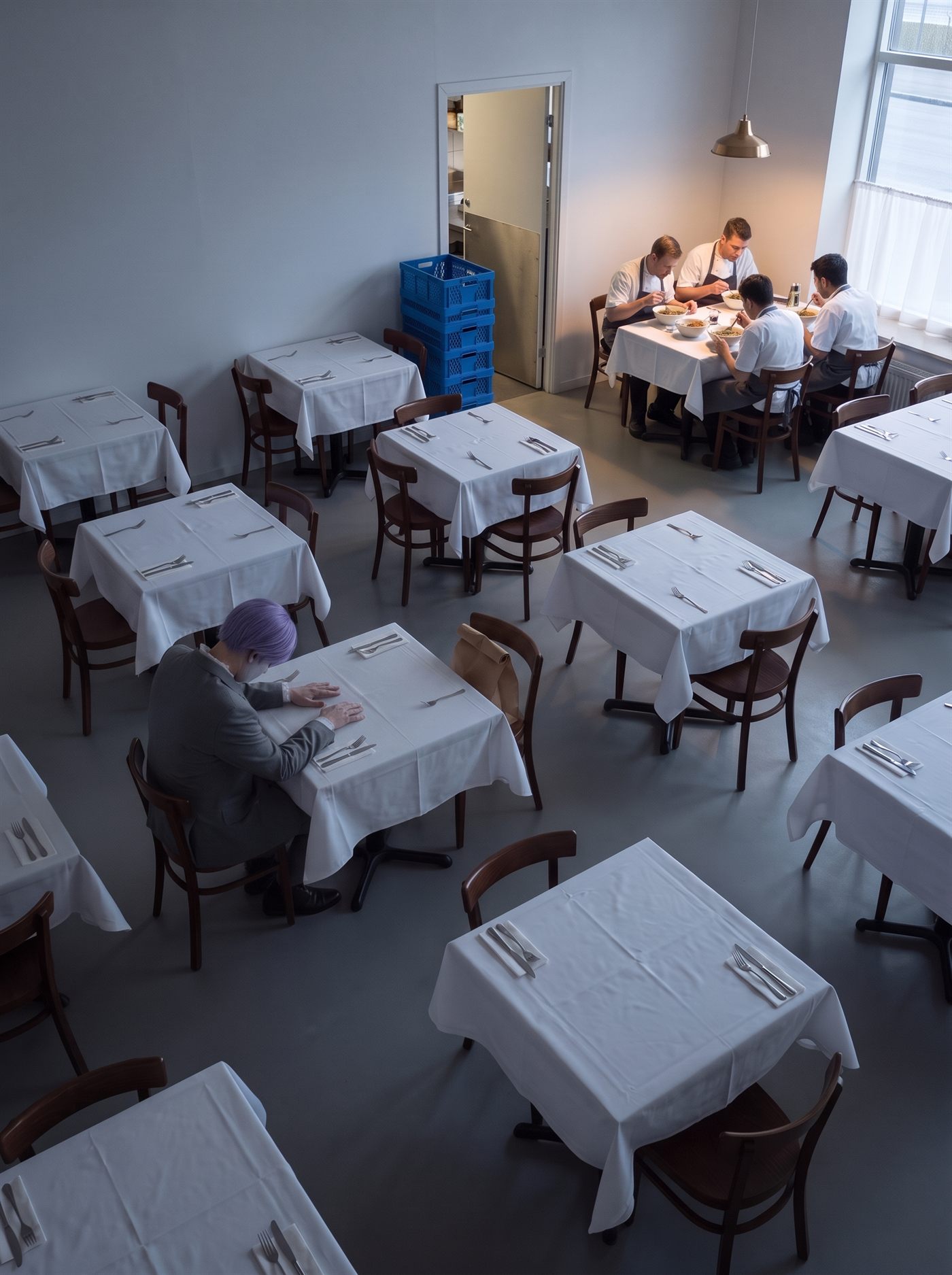 A photograph taken from high in the corner of a small restaurant dining room, looking down across it in the late afternoon. Fifteen or more square tables stand in loose rows under plain white cloths, each laid with cutlery, every chair pushed in, every table empty; the room is lit only by cold flat daylight from a tall window on the right and the floor between the tables is a dark grey. At one table in the lower left, small and well off-centre, a man in a grey suit half a size too big sits alone with his head bowed straight down over an empty place setting, both hands laid flat on the cloth. He is seen from directly behind and above, so nothing of him shows but the crown of his short lavender hair, his shoulders and his hands. A small brown paper bag with its top rolled over once stands upright on the seat of the empty chair beside him. Four or five empty tables away, at the far end of the room under the only lamp that is switched on, four restaurant workers in aprons sit close together at two tables pushed into one, eating out of big steel bowls and leaning in toward each other. None of them is looking at him. Beside them, in the open kitchen doorway, a stack of blue plastic delivery crates is the only strong colour in the room.