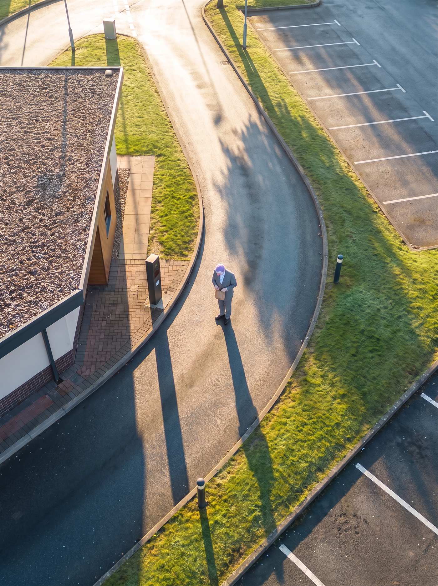A photograph taken from high above a small fast-food restaurant just after sunrise, looking steeply down. On the left is the corner of the building, its flat roof covered in brown gravel and a paved brick path running along its side past a short dark order post. A single curving lane of dark asphalt, still wet, sweeps around the building from the bottom of the frame toward the top, edged by low concrete kerbs and strips of grass silvered with dew; beyond it on the right are two empty car parks marked with plain white bay lines. There are no cars anywhere. The sun is very low, and everything throws long shadows the same way: the bollards, the light poles, the kerbs. In the middle of the lane, beside the order post, where a car's window would be, stands one small man on foot in an off-the-rack grey suit, a white collar at his neck and a lanyard hanging down his shirt. His head is bowed over a small brown paper bag held in both hands at his waist, so that from above we see mostly his short lavender hair, a white-painted face and the round pink tip of a clown's ball nose. His shadow stretches a long way back up the lane behind him. Nobody else is in the picture.