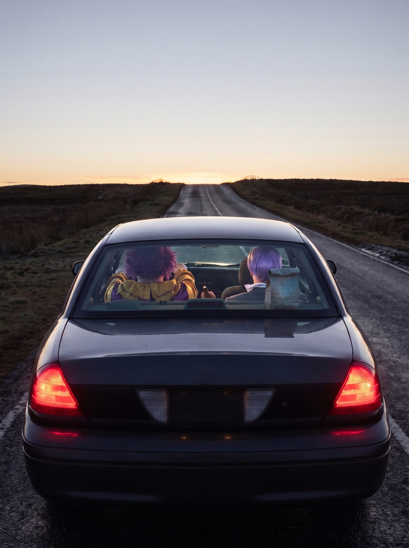 A photograph taken from a few paces directly behind a large dark sedan standing still in the middle of an empty two-lane road, which runs dead straight away from the camera across open moorland to a low crest on the horizon. The sun has not risen. The sky above the crest is a broad band of pale gold going up into cold grey-blue, and the light is coming from somewhere beyond the top of the rise, lighting the underside of the sky and nothing else; the moor on either side is dark and colourless. The car's rear lights are lit red and its number-plate recess is empty and unmarked. Through the rear glass both occupants are seen squarely from behind, the backs of their heads to the camera. In the driver's seat on the left a heavyset figure is slumped forward over the steering wheel, a wild aubergine-purple mop of hair blown in every direction above a tattered mustard-yellow ruffled collar and purple sleeves. In the front passenger seat on the right a man sits upright and very still, the back of his head to the camera: a short lavender wig combed flat above a small white ruffled collar and the grey shoulders of a suit, his head level, facing the top of the hill. Between the two seats a hand rests on a handbrake lever standing pulled all the way up. On the parcel shelf under the rear glass, beside the man's shoulder, stands a small plain brown paper bag with its top rolled shut and no handles. The road ahead of the car is empty the whole way to the crest, and nothing on the other side of it can be seen.